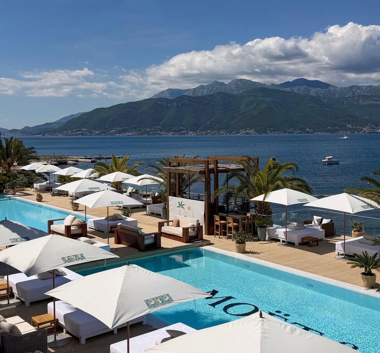 Bazen Monte Palm Beach s bijelim ležaljkama i suncobranima, zaliv i planine iznad Tivta