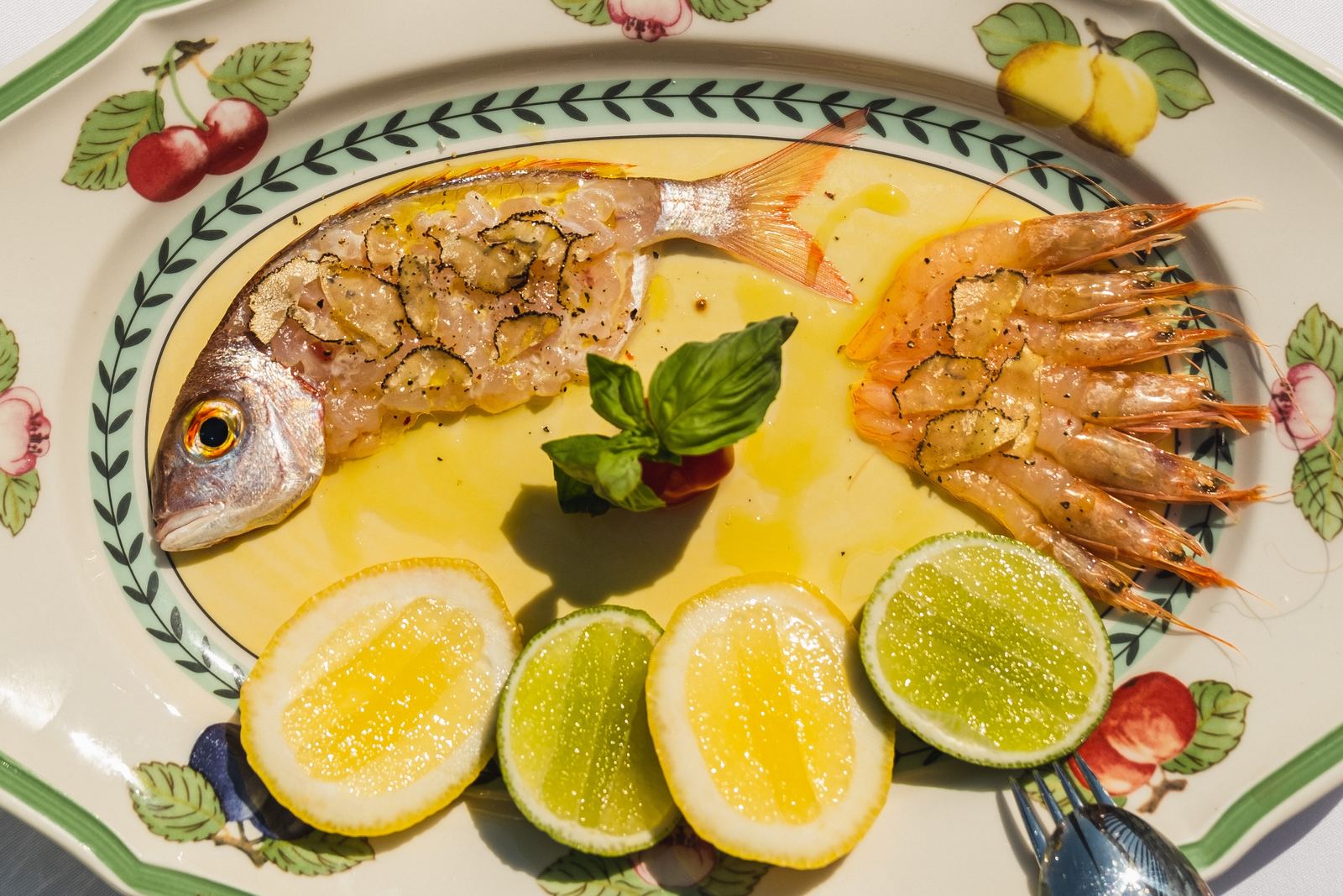 Cijela riba i škampi na oslikanom tanjiru s limunom i limetom, restoran Špilja u Luštica Bayu
