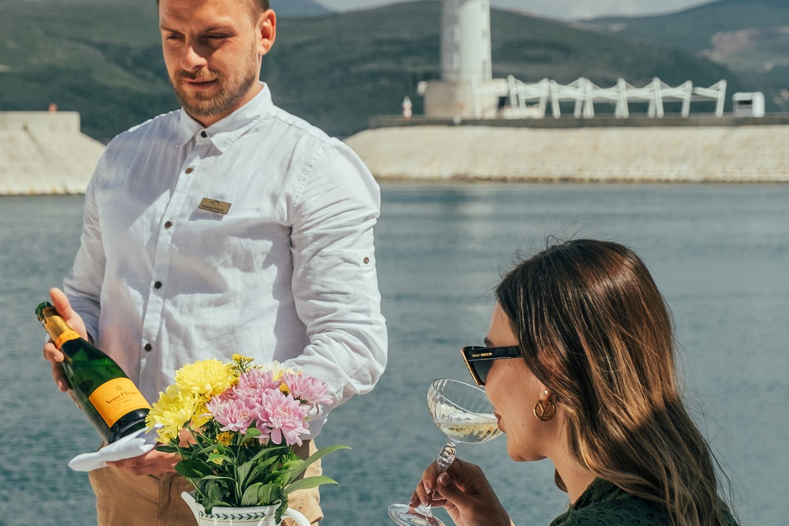 Konobar restorana Špilja služi šampanjac gošći za stolom uz more, svjetionik Luštica Baya iza